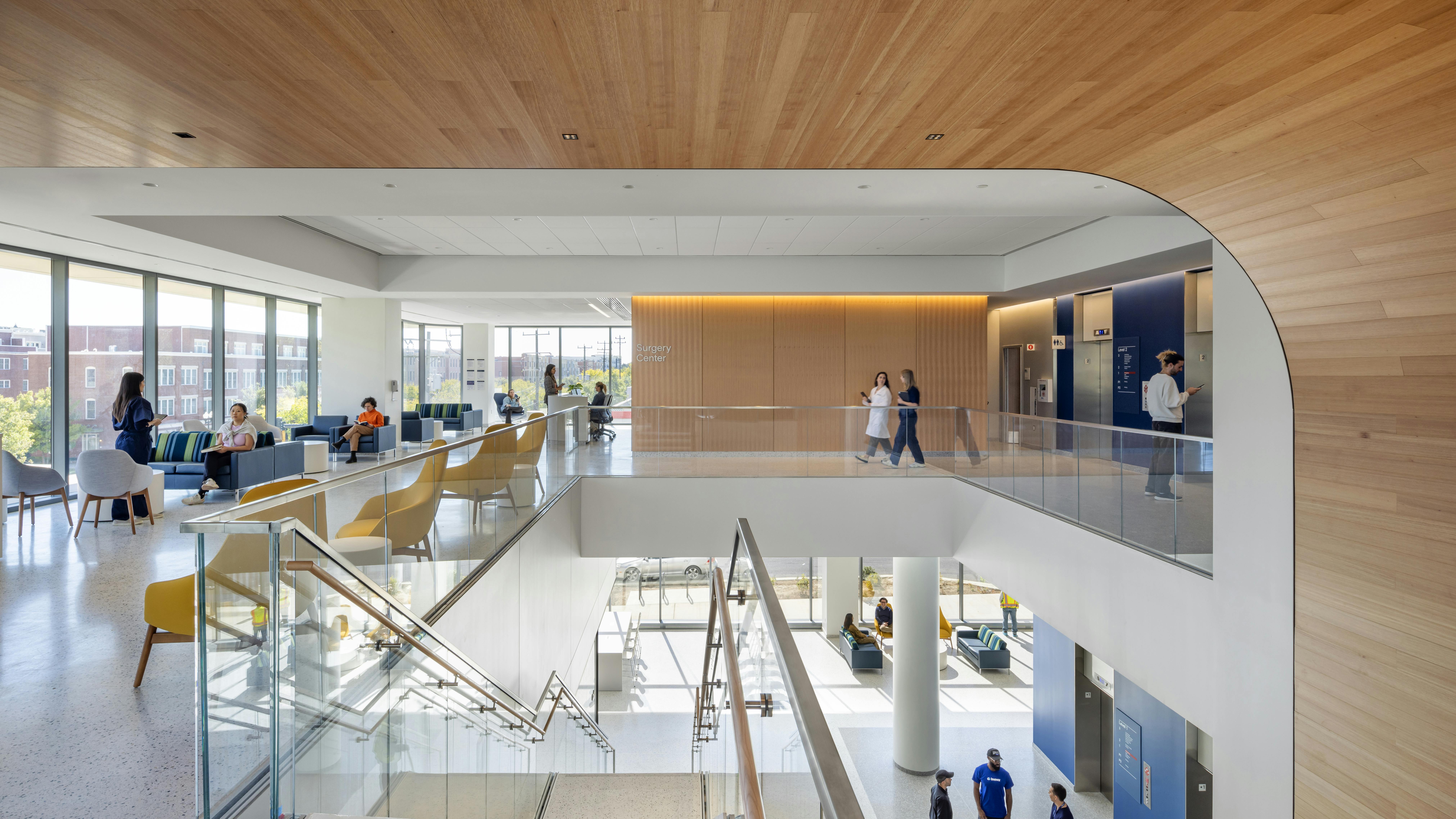Interior view from top of staircase in two-story Inova Health Center featuring a curved, wood wall, seating areas next to large daylit windows and people interacting in the space.