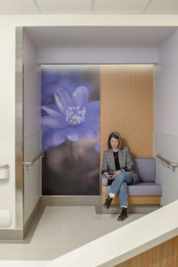 Woman sitting on upholstered bench in small hospital alcove with wood paneling and botanical wall graphic. Woman sitting on upholstered bench in small hospital alcove with wood paneling and botanical wall graphic.