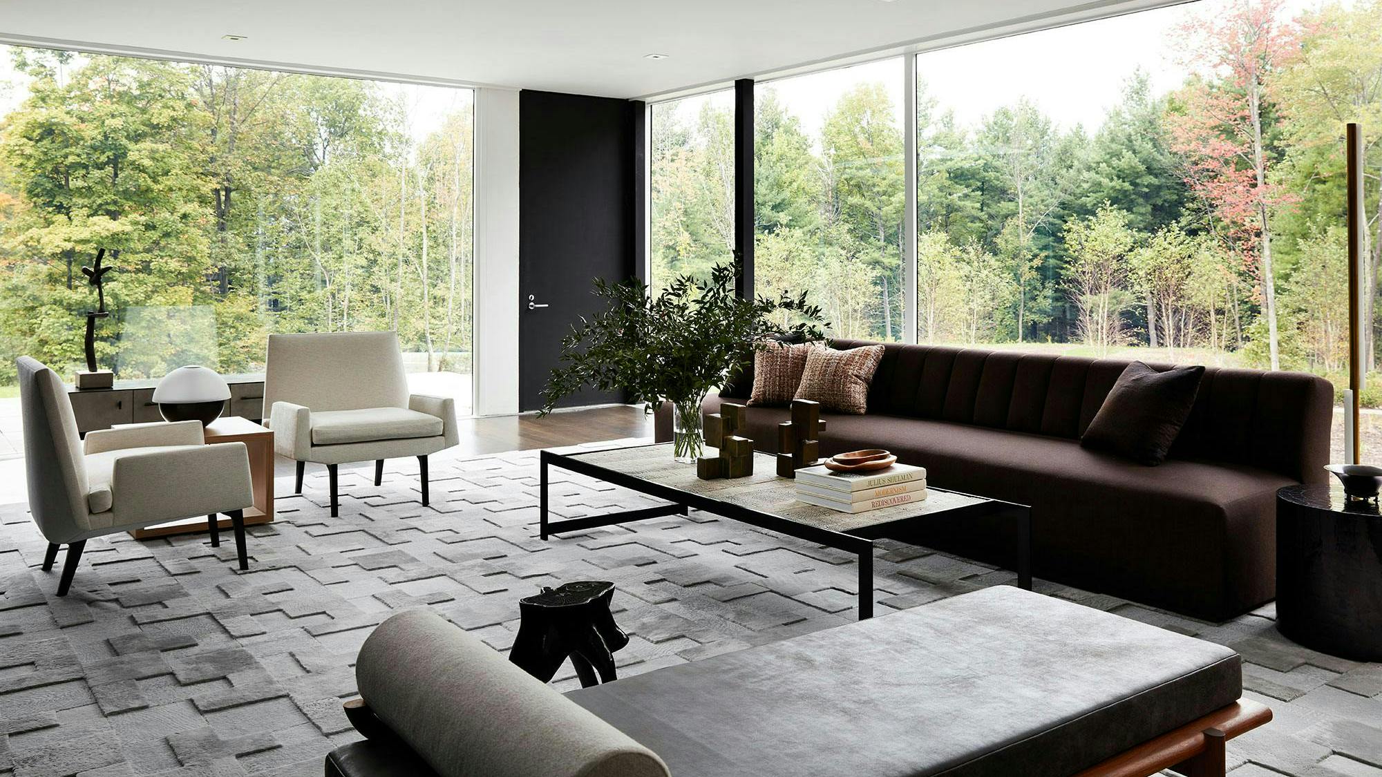 A modern sitting room with expansive window views to the surrounding treeline, with foliage changing in the fall