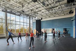 Dance studio space with dark rubber flooring and several female students posing with large glass wall in background. Dance studio space with dark rubber flooring and several female students posing with large glass wall in background.
