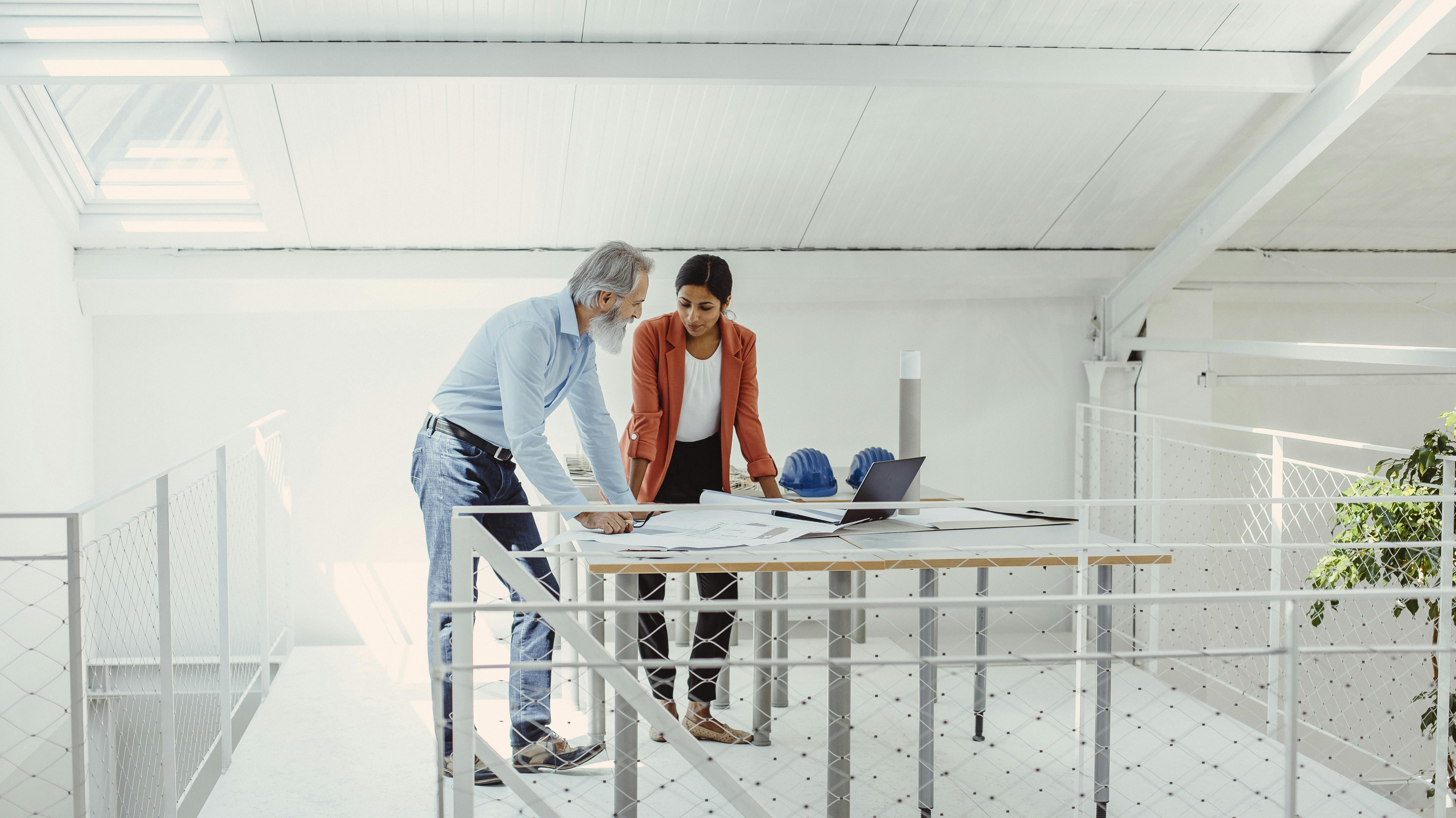 Older man talking with younger woman in mentorship role looking over blueprints in bright office.