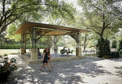 Open pavilion constructed of steel, wood, and stone with seated visitors, with gardens and greenery in the background. Open pavilion constructed of steel, wood, and stone with seated visitors, with gardens and greenery in the background.