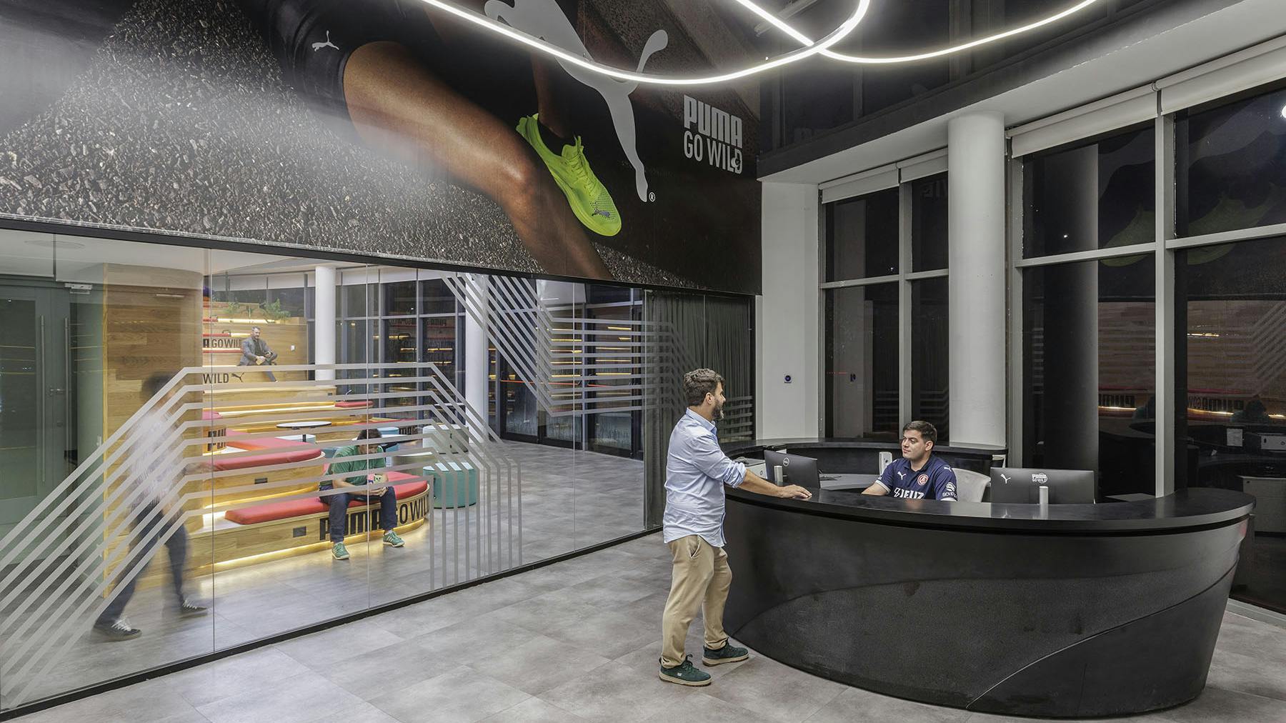 Puma corporate employees at circular stone reception desk with large glass-walled view to tiered stadium-bench style seating.