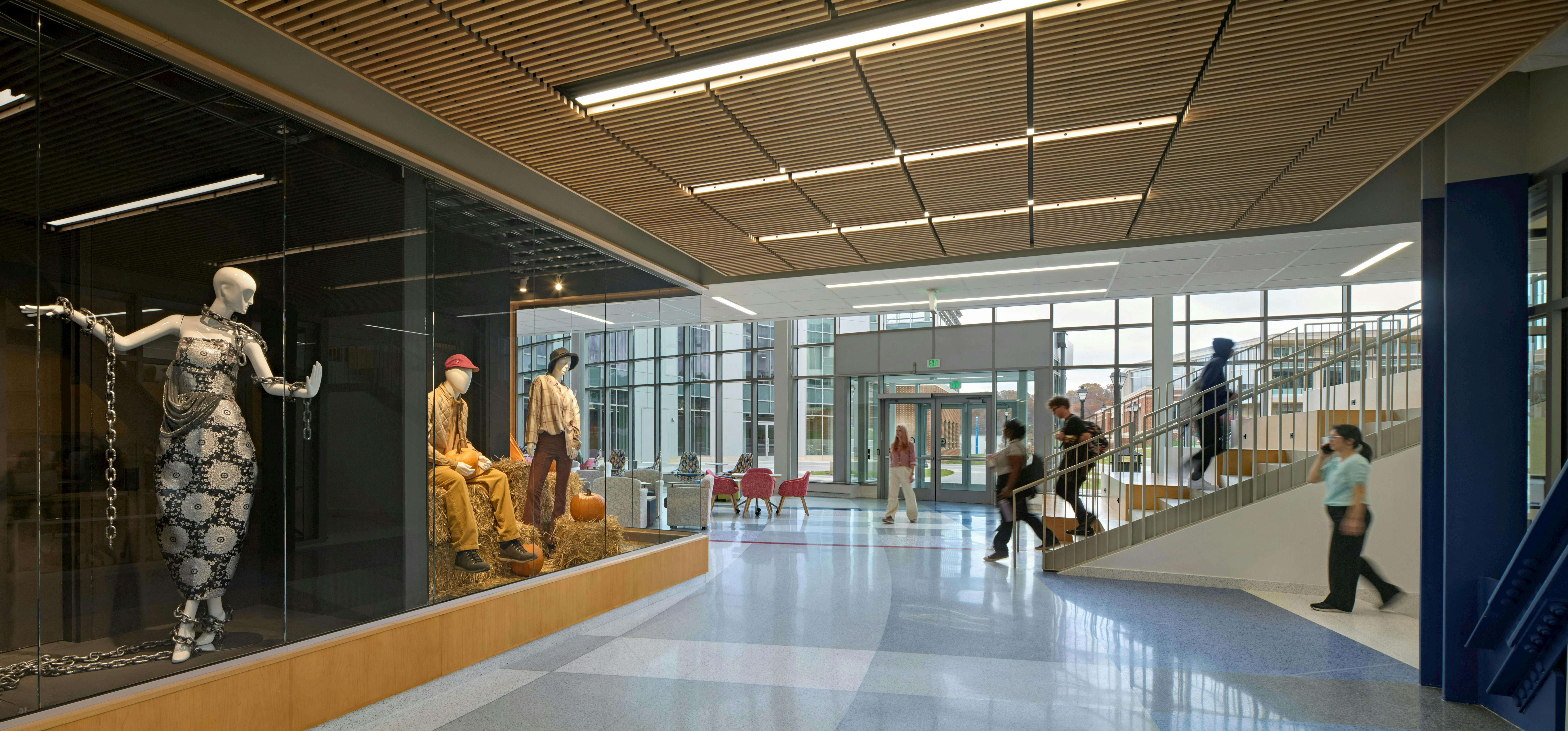 Hallway with glass wall showing fashion mannequins in foreground with staircase and glass wall with students walking in background.