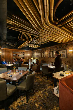 Exposed ceiling in the lounge features gold-painted pipes overhead, and dark-toned furnishings in multiple seating arrangements, with sumptuous leather and velvet textiles. Exposed ceiling in the lounge features gold-painted pipes overhead, and dark-toned furnishings in multiple seating arrangements, with sumptuous leather and velvet textiles.