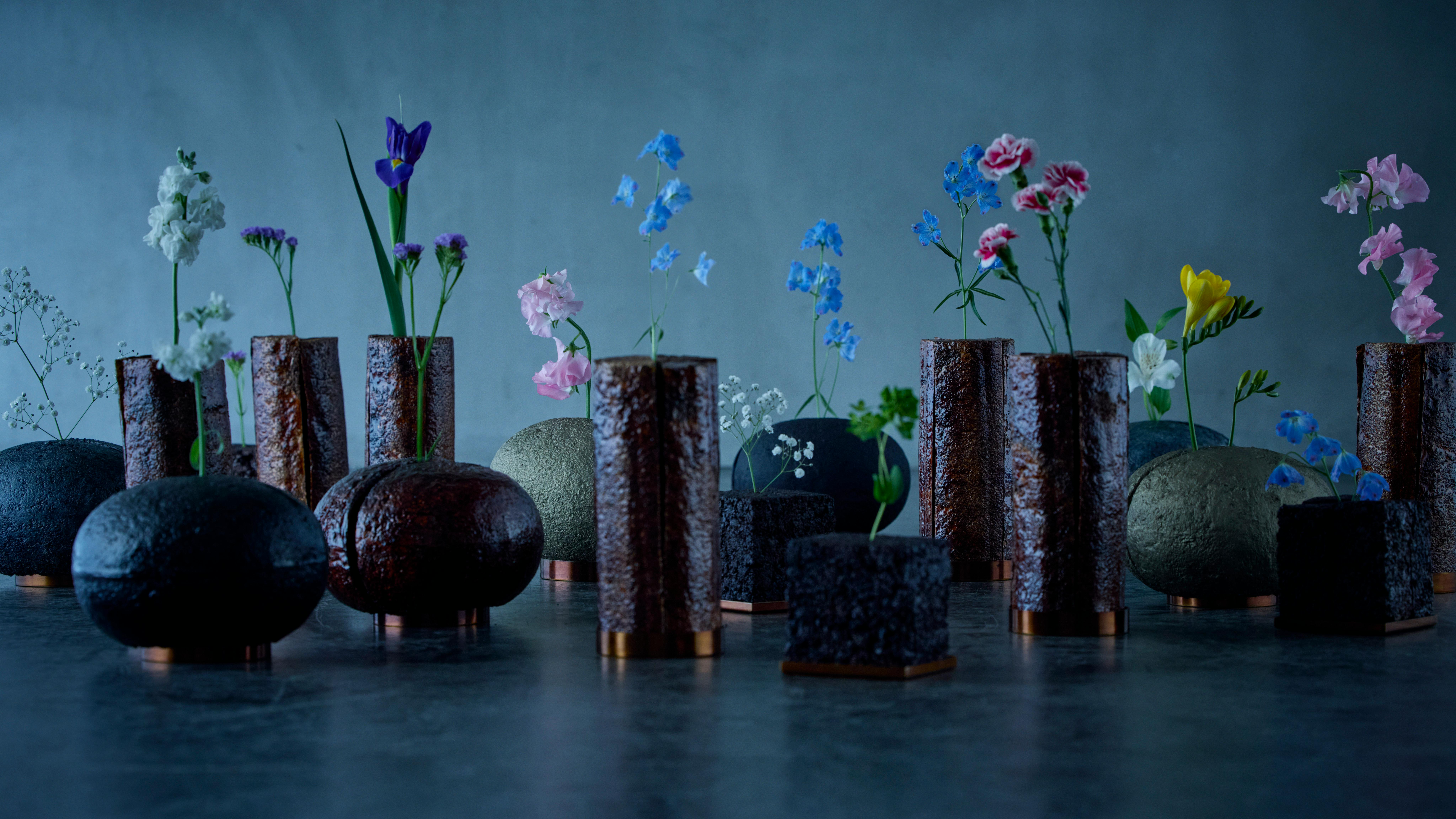 A variety of dark-colored vases with flowers against a blue background.