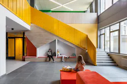 Bright yellow staircase with students in cubbies underneath in background and soft, red seating area with one student in foreground. Bright yellow staircase with students in cubbies underneath in background and soft, red seating area with one student in foreground.