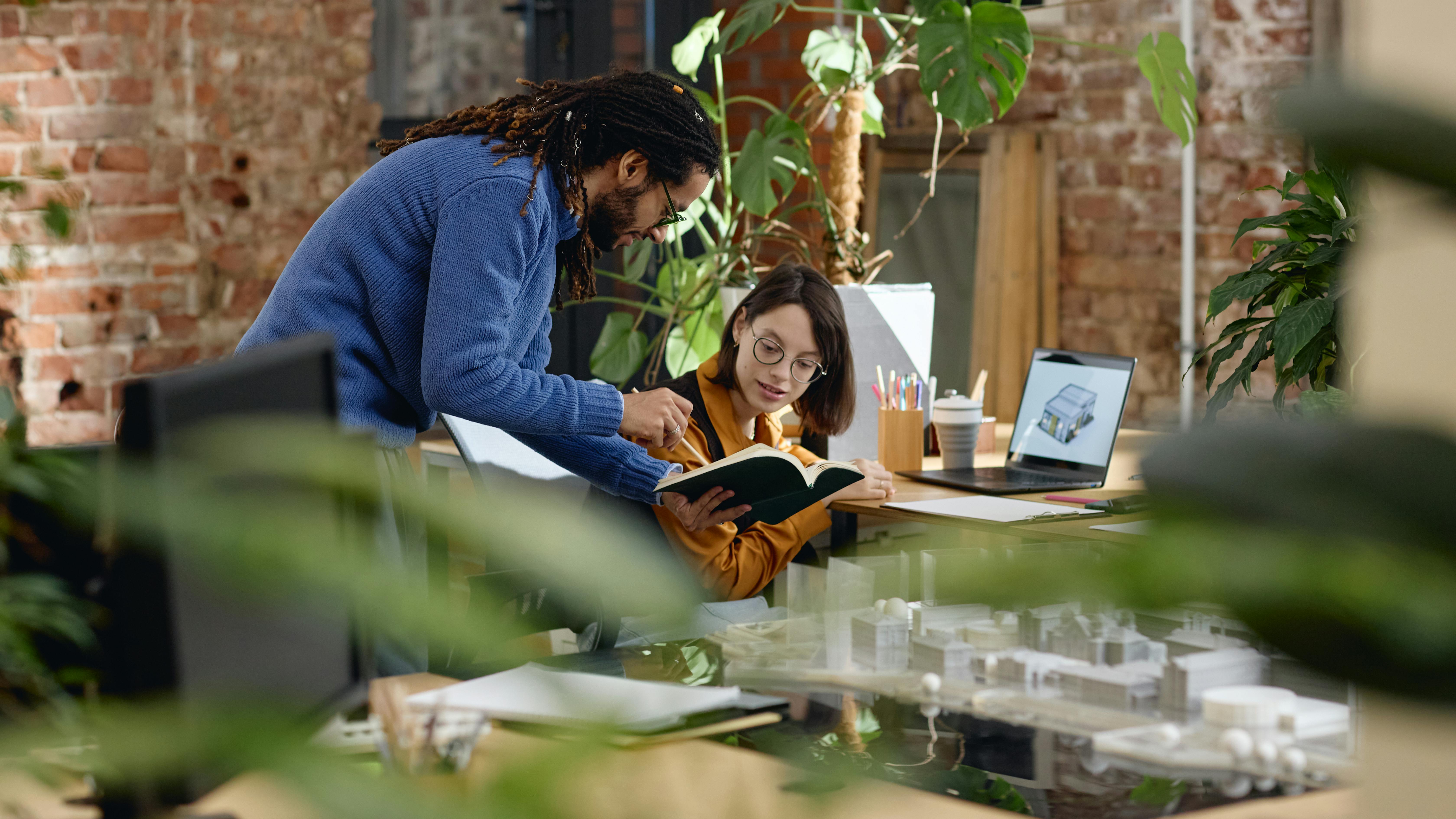 Black man standing beside seated Caucasian young adult woman, both collaborating over open notebook at modern office desk, architectural models and laptop visible on workspace