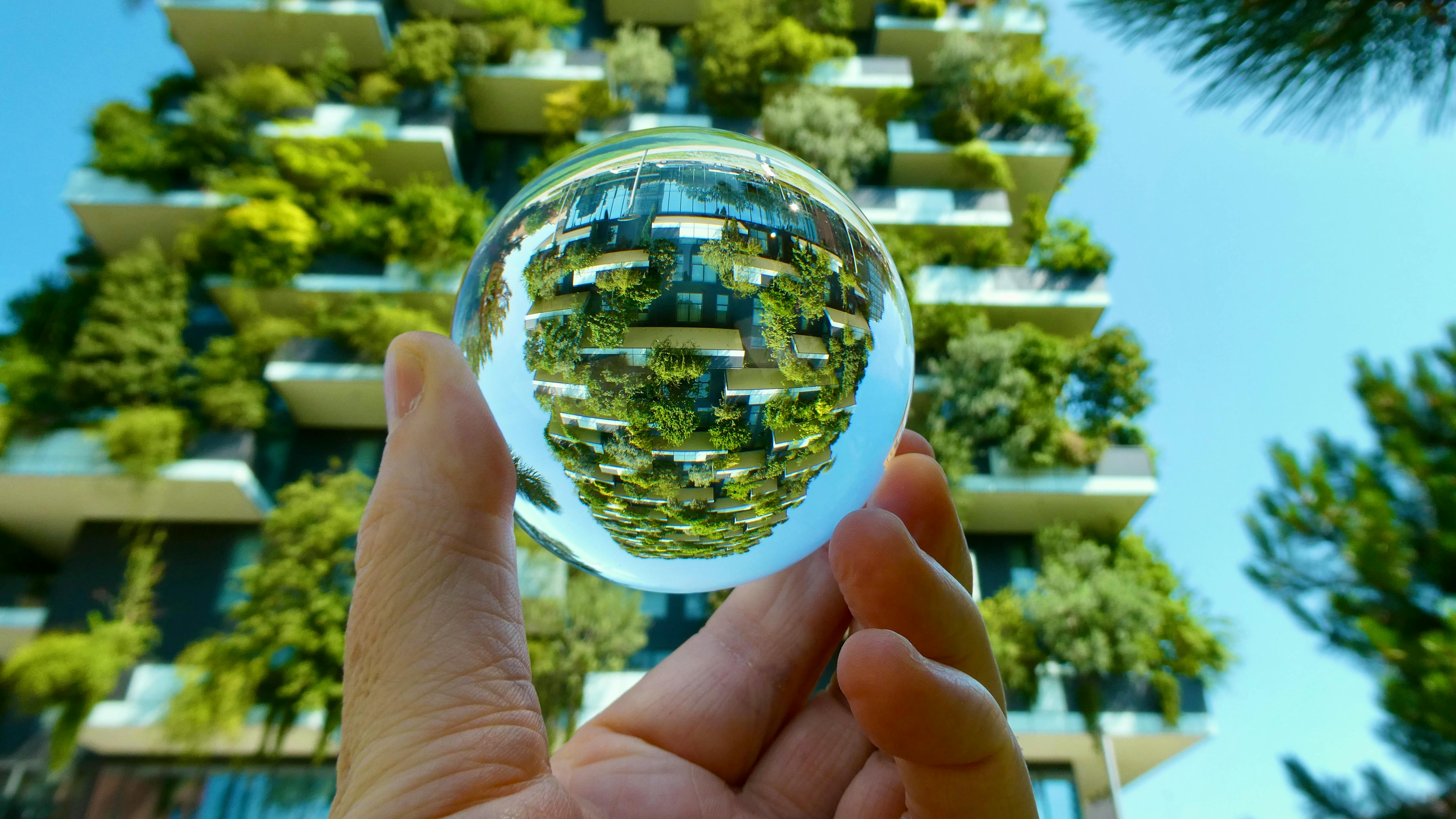 Hand holding a glass orb with tall building covered in green plants.