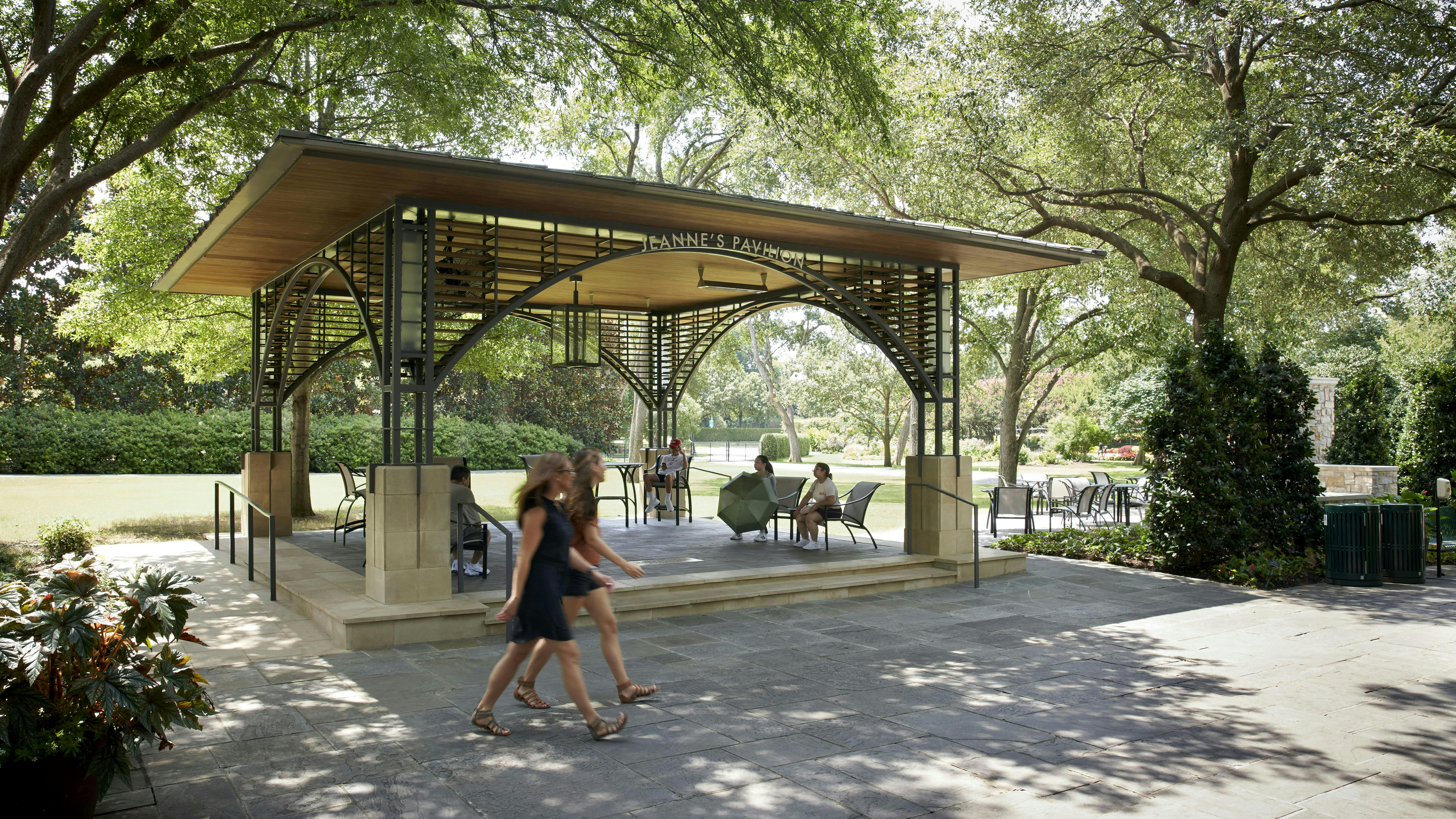 Open pavilion constructed of steel, wood, and stone with seated visitors, with gardens and greenery in the background.
