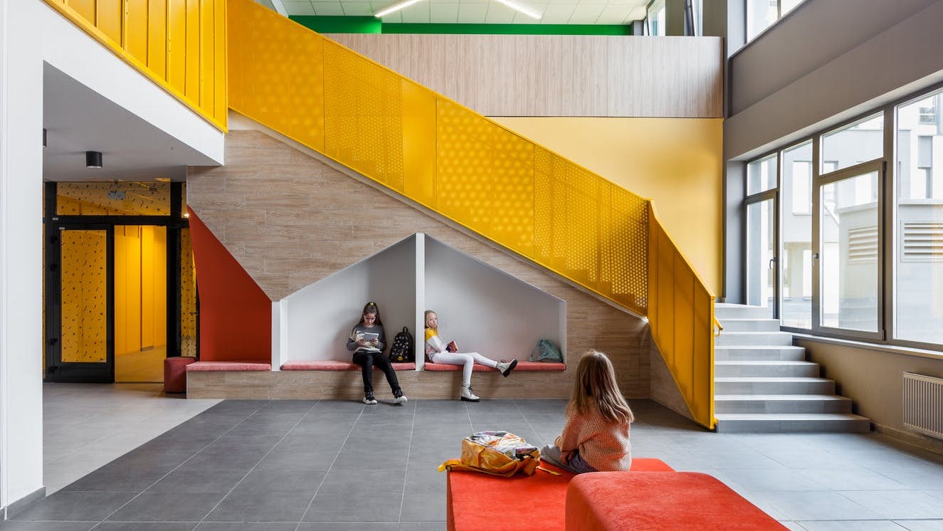 Open communal space in school with geometric shaped seating alcoves, soft lounge seating, and bright yellow staircase