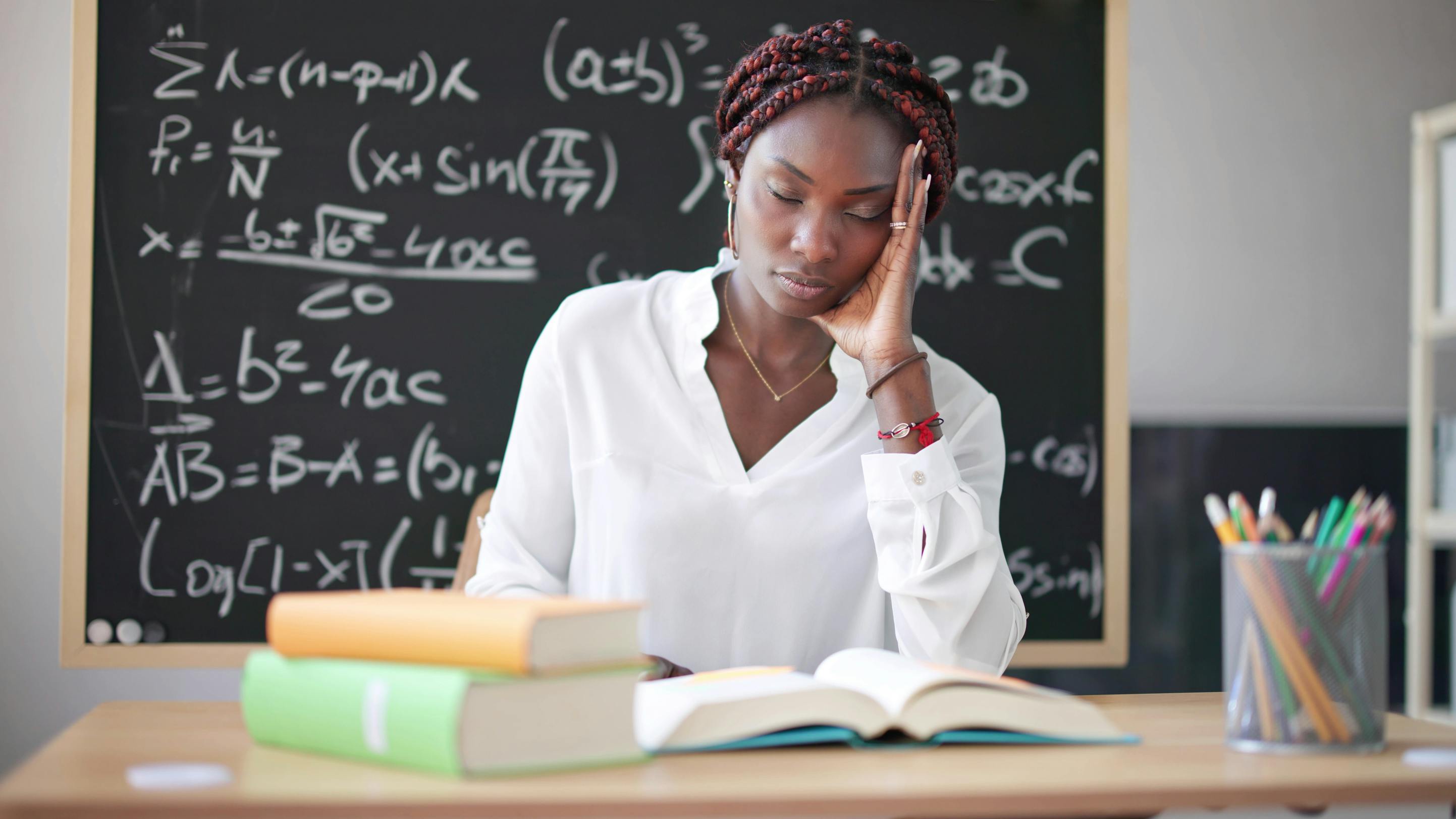 A young Black female teacher sits at a desk leaning her hand on her hand