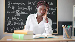 A young Black female teacher sits at a desk leaning her hand on her hand