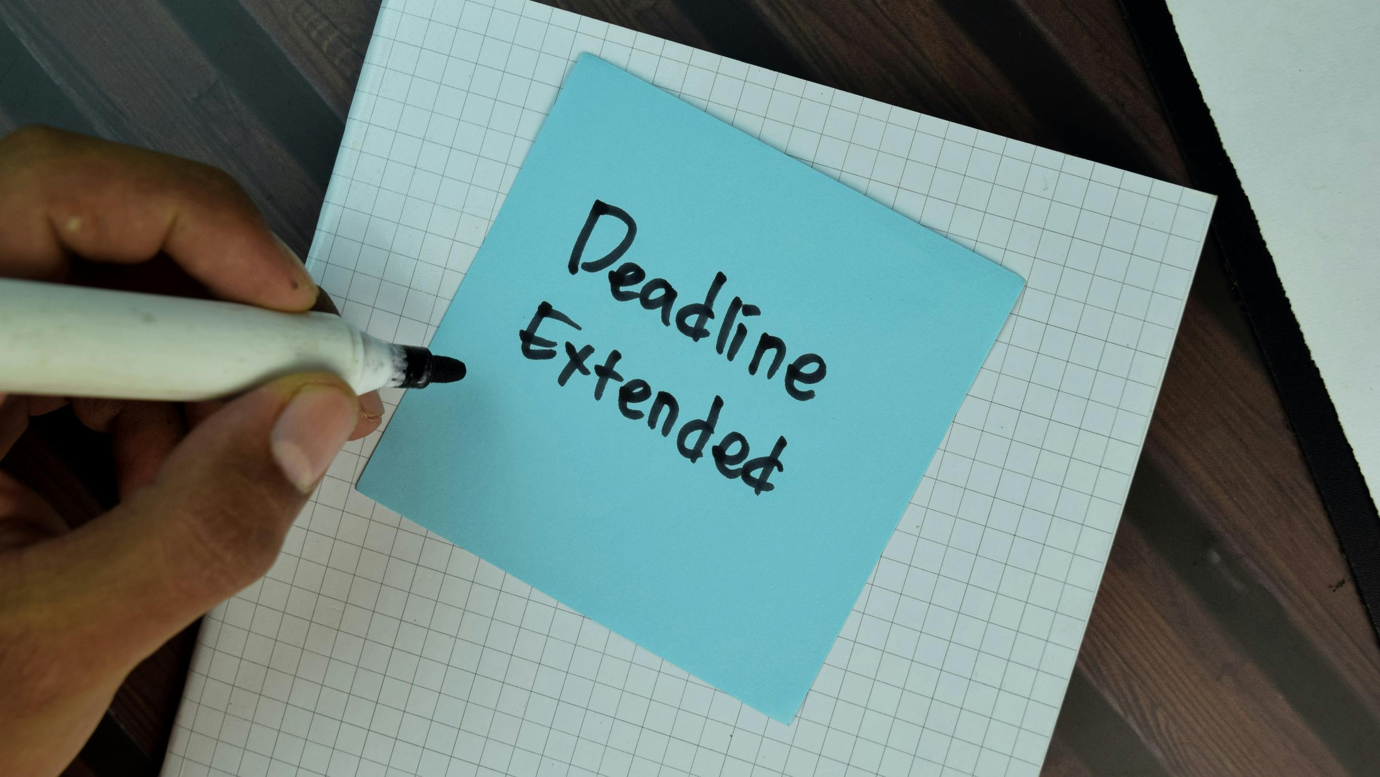 Off frame person writes Deadline Extended on a blue sticky note sitting on top of graph paper