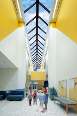 An airy ambiance is achieved under the extensive skylight, with white walls and speckled flooring anchoring the yellow interior stairwell and blue furnishings.