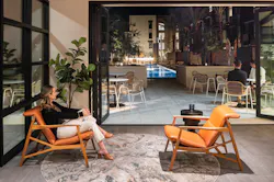 A woman sits in an orange leather-upholstered lounge chair inside a seating area adjacent to the pool lounge, with glass doors open to the evening air