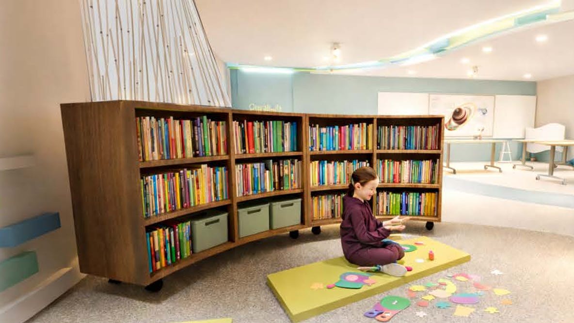 Design rendering shows a female elementary school student playing with shapes in a quiet area of a classroom set off by a curved bookcase and partial curtain