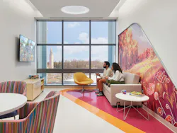 Man and woman sit in a family room at hospital, decorated with shades of pink, purple, green, blue, and orange tempered with simple and neutral wood doors and furnishings, white surfaces, and layered white and color accent lighting.