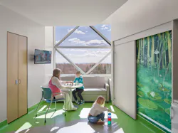 Woman seated at a round table plays a game with young boy, while young girl p[ays with toys on the floor in a bright, sunlight-filled room with a green mural on the wall.