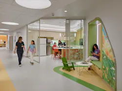 Medical staff person walks the hall with a young girl, while a young woman reads in a seating nook along the wall. A woman and young boy are in the kitchen in the background.