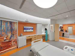 Medical worker stands at a sink in a large patient room outfitted in vibrant orange accents that match the hall wayfinding color.