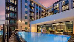 Man and woman sit on the edge of outdoor infinity pool adjacent to a large, windowed community room at a multiresidential community.