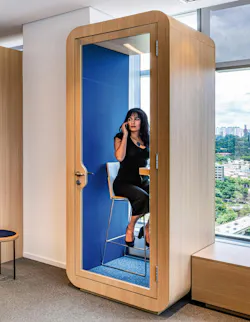 A woman sits in an enclosed, single-occupant privacy booth with glass door, blue interior walls, and light wood exterior materials
