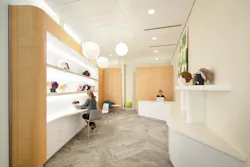 A woman sits in front of lighted display shelves holding wigs, while a female receptionist works on a computer in the background