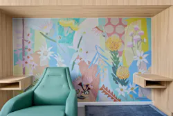 A lounge nook with light wood inspired built-ins and surfaces, a curved turquoise lounger, and botanical wall mural in soft gold, lavender, green, blue, and peach tones