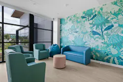 A common room features a bold floral wall mural anchored with green, blue, and neutral tones reflected in the soft seating and views to the exterior landscape