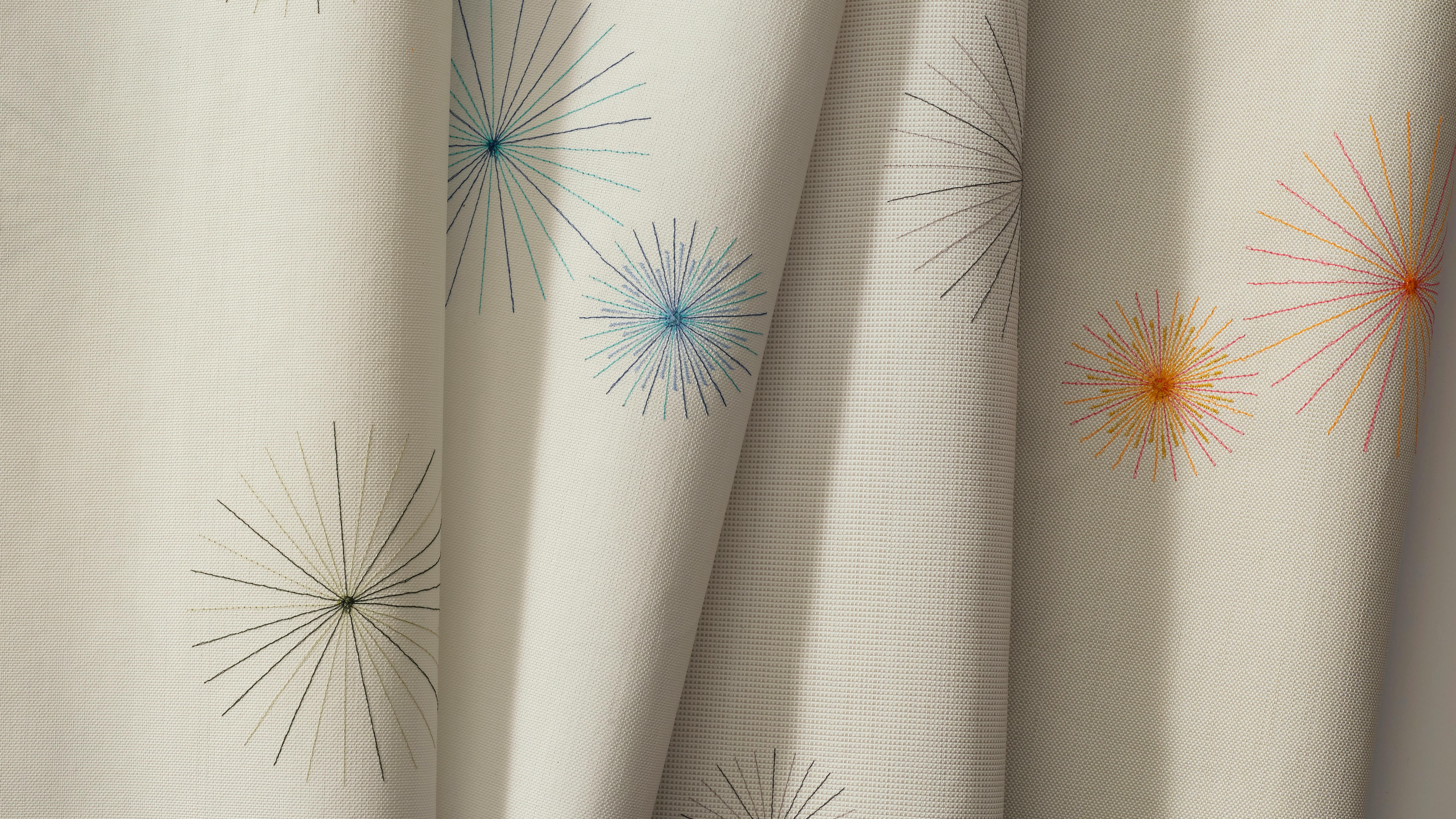 Aperture Embroider textile features finely stitched radial bursts across an open ground. Shown with black, blue, and orange bursts.