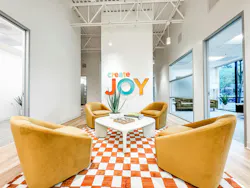 White-walled lounge with multicolor wall sign that says Create Joy, golden yellow club chairs, and a deep orange-and-white checked area rug