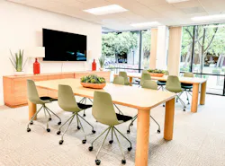 A group meeting room with light tables and credenzas, green wheeled task chairs, and large windows showing a courtyard