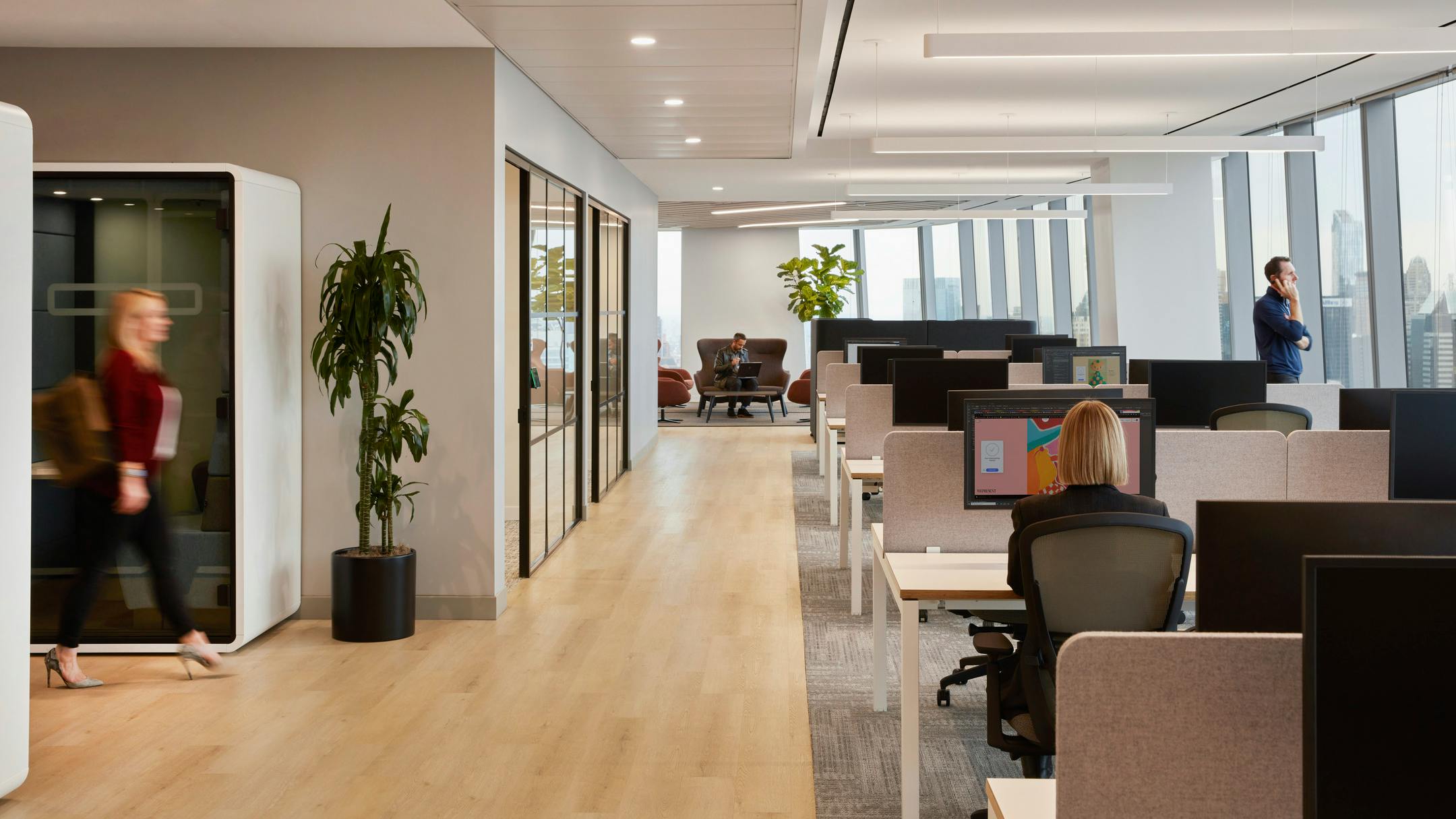 An interior workplace with individual privacy booths, glass-walled conference room, open desk zone with low acoustic barriers, and a casual seating area at the far end of the room