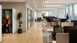 An interior workplace with individual privacy booths, glass-walled conference room, open desk zone with low acoustic barriers, and a casual seating area at the far end of the room