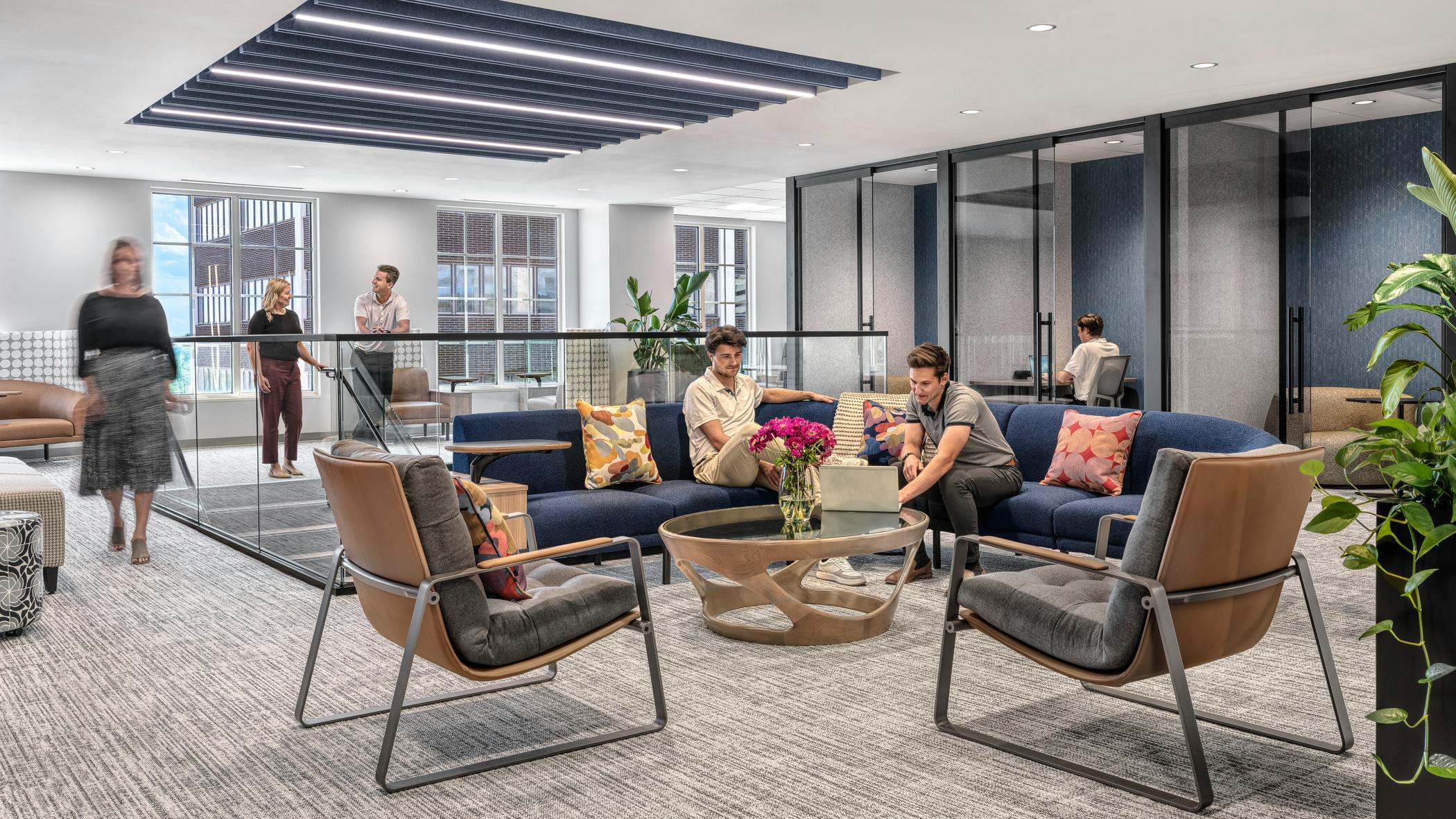 Several employees gather in an open seating area on a curved, blue-upholstered modular sofa, as others converse atop a glassed-in staircase nearby
