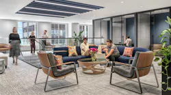 Several employees gather in an open seating area on a curved, blue-upholstered modular sofa, as others converse atop a glassed-in staircase nearby