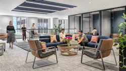 Several employees gather in an open seating area on a curved, blue-upholstered modular sofa, as others converse atop a glassed-in staircase nearby