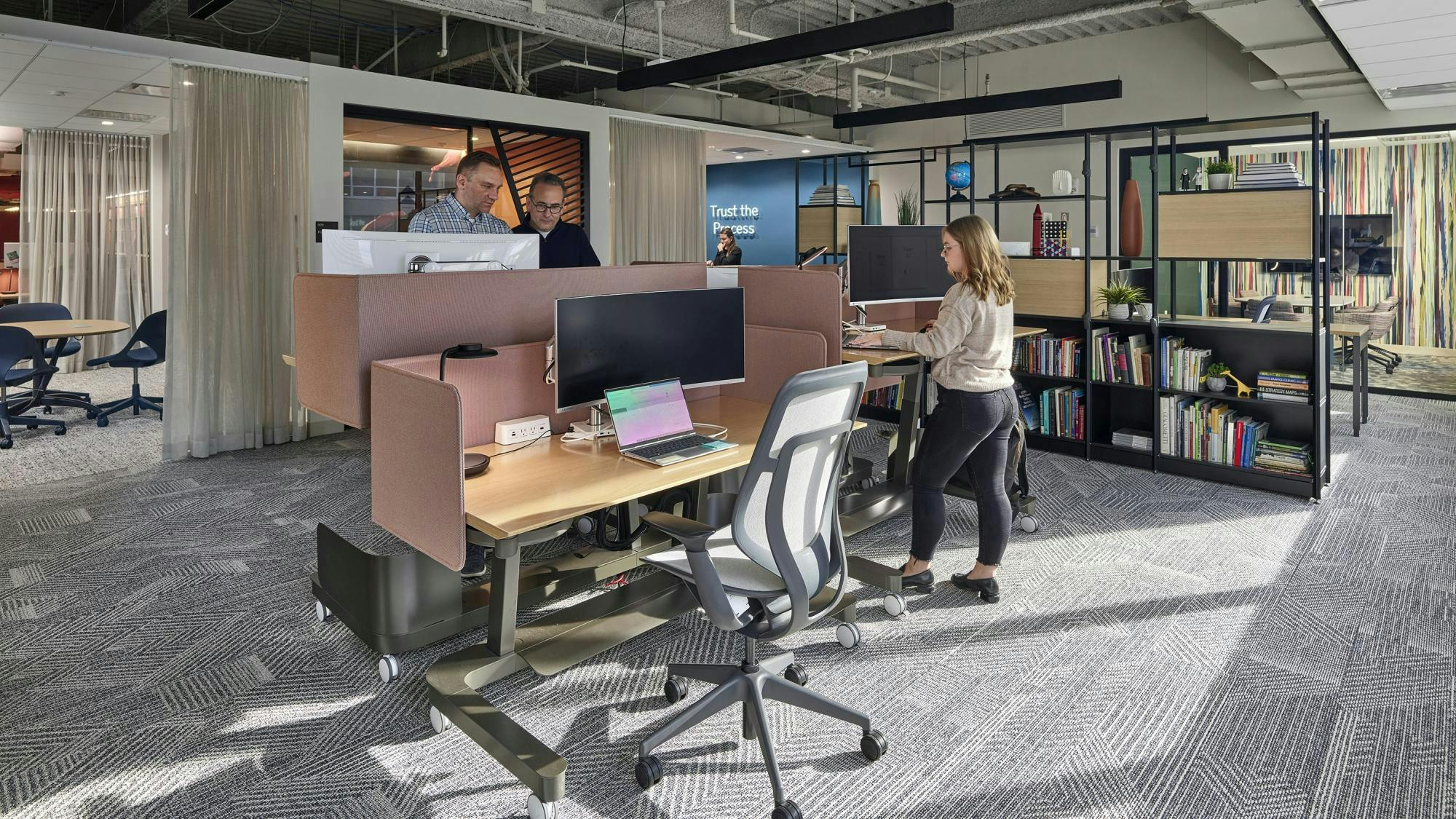 Employees work at sit-stand desks with acoustic screens in an open office area