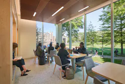 Students relax or study in various group and individual seating arrangements in a lounge with large campus-facing windows
