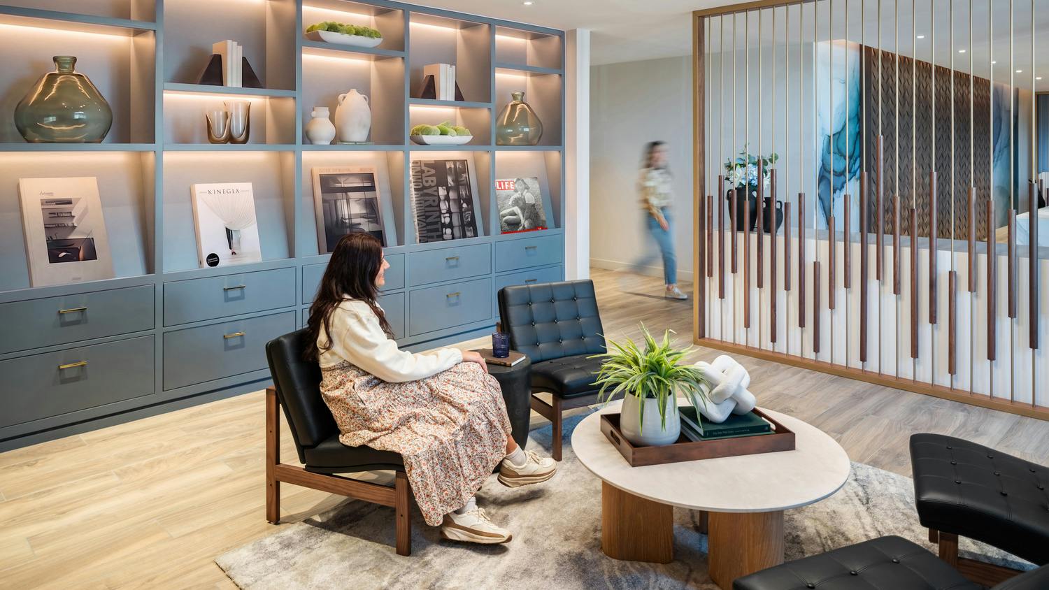 A woman sits in a wood-frame upholstered chair in a private lounge space featuring a built-in wall display for decor and accessories and a decorative divider