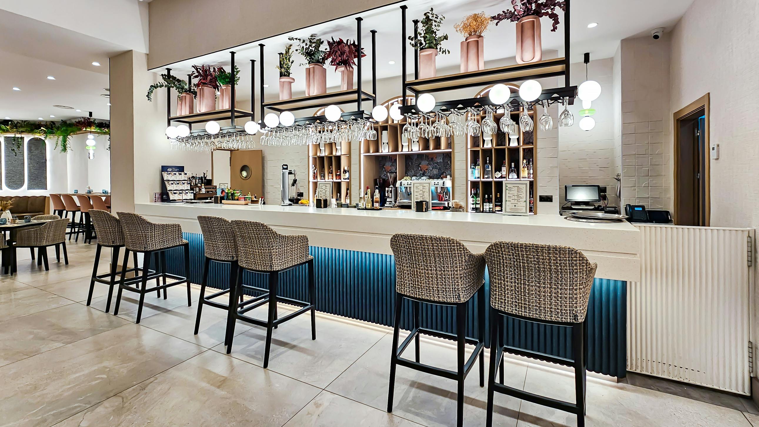 Contemporary and cozy hotel bar with high-back bar-height seating, light neutral stone and tile surfaces, ceiling-mounted shelves combined with globe light fixtures and faux floral arrangements