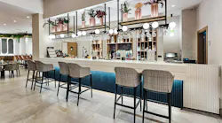Contemporary and cozy hotel bar with high-back bar-height seating, light neutral stone and tile surfaces, ceiling-mounted shelves combined with globe light fixtures and faux floral arrangements