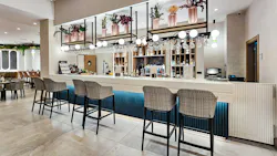 Contemporary and cozy hotel bar with high-back bar-height seating, light neutral stone and tile surfaces, ceiling-mounted shelves combined with globe light fixtures and faux floral arrangements