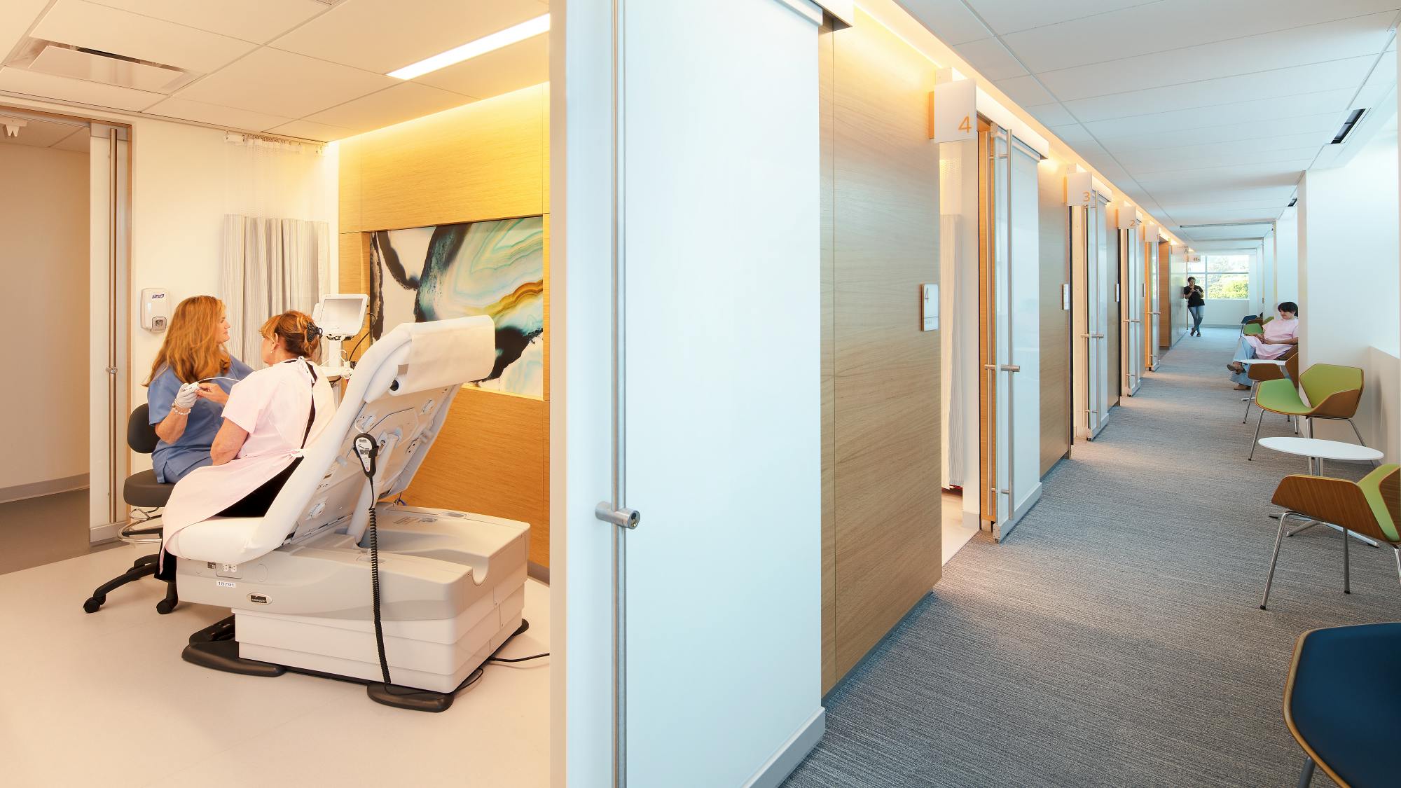 Female healthcare provider examines a female patient in a comfortable exam room, with a light-filled corridor adjacent